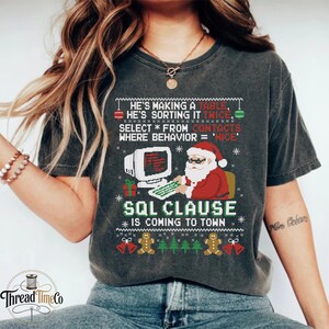 May include: A charcoal gray t-shirt featuring a Christmas-themed graphic with Santa Claus at a computer. The text reads: HE'S MAKING A TABLE, HE'S SORTING IT TWICE, SELECT * FROM CONTACTS WHERE BEHAVIOR = 'NICE', SQL CLAUSE IS COMING TO TOWN.