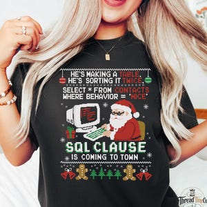 May include: A black t-shirt featuring a pixelated graphic of Santa Claus at a computer. The text reads: He's making a table, he's sorting it twice, select * from contacts where behavior = 'nice', SQL clause is coming to town, with gingerbread men.