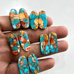 Könnte beinhalten: Eine Sammlung von acht polierten Cabochons mit einem Mosaikmuster aus leuchtend türkisfarbenen, kräftig orangefarbenen und weißen Steinfragmenten. Die Steine sind oval und rechteckig geformt und zeigen eine marmorierte Textur mit metallischen Adern.