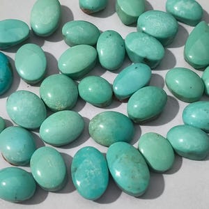 Könnte beinhalten: Eine Sammlung loser, polierter Türkis-Edelsteine in verschiedenen ovalen und runden Formen. Die Steine zeigen eine Reihe lebendiger blau-grüner Farbtöne mit glatten, gewölbten Oberflächen, natürlichen Farbvariationen und subtilen Matrixmustern.