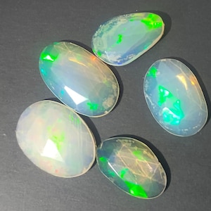 Könnte beinhalten: Fünf facettierte Opale in ovaler Form mit einer durchscheinenden, milchig-weißen Grundfarbe. Jeder Edelstein zeigt lebhafte neon-grüne Blitze und ein subtiles orangefarbenes Lichtspiel auf den polierten, facettierten Oberflächen.