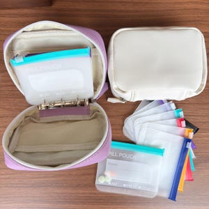 May include: A lavender zippered organizer case with a metal ring binder mechanism inside, accompanied by a white zippered case and several translucent plastic pouches labeled PILL POUCH with fields for name, day, and time settings of AM, PM, or other.