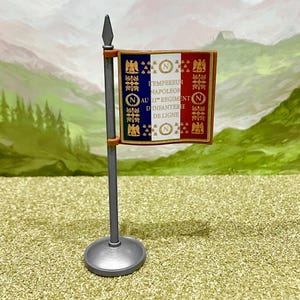 Könnte beinhalten: Eine Miniaturflagge an einem grauen Mast mit rundem Sockel. Die Flagge zeigt vertikale blaue, weiße und rote Streifen mit goldenen Adler- und N-Emblemen. Der Text lautet: L'EMPEREUR NAPOLEON AU 21E REGIMENT D'INFANTERIE DE LIGNE mit N-Symbolen.