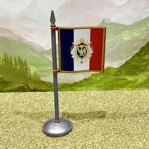 Könnte beinhalten: Eine französische Miniaturflagge an einem silberfarbenen Mast mit rundem Sockel. Die Flagge zeigt vertikale blaue, weiße und rote Streifen mit einem zentralen goldenen kaiserlichen Adleremblem, einer Krone und verzierten Details auf dem weißen Streifen.