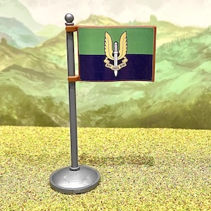 May include: A miniature desk flag featuring a horizontal bicolor design of light green and dark blue. Centered on the flag is a gold winged dagger emblem with a scroll underneath displaying the text Who Dares Wins. The flag is attached to a gray pole.