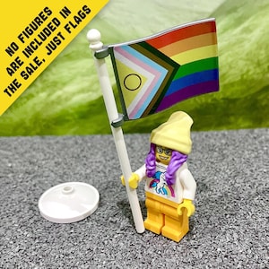 Op de afbeelding: Een plastic speelgoedvlag met het Progress Pride-ontwerp met horizontale regenboogstrepen en een chevronpatroon met witte, roze, lichtblauwe, bruine en zwarte kleuren en een paarse cirkel. De tekst luidt: NO FIGURES ARE INCLUDED IN THE SALE, JUST FLAGS.