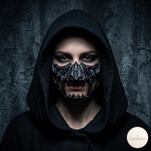 Könnte beinhalten: Eine schwarze, texturierte und skulptierte Gesichtsmaske mit scharfen, fangzahnartigen Vorsprüngen und einem gezackten, skelettartigen Design. Die Maske bedeckt die untere Gesichtshälfte von der Nase bis zum Kinn und wird mit einer schwarzen Kapuze getragen.