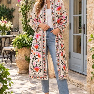 May include: A long, cream-colored open-front duster coat featuring vibrant floral embroidery in shades of pink, orange, and black. The garment is worn over a plain white top and light blue denim jeans with a straight-leg cut and raw hem.