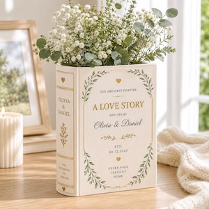 May include: A cream-colored book-shaped vase featuring gold text that reads A LOVE STORY, WRITTEN BY Olivia & Daniel, EST. 04.12.2025, EVERY PAGE LEADS US HOME, and OUR GREATEST CHAPTER, decorated with green leaf borders and small gold hearts.