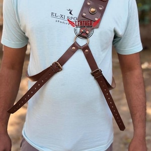 May include: A brown leather chest harness featuring a wide shoulder strap adorned with metallic studs and a central brass ring. The harness includes adjustable straps with buckles and metal rivets, worn over a light blue t-shirt with printed text.