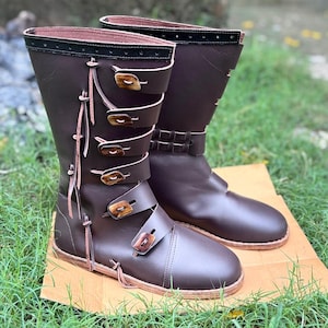 May include: A pair of dark brown leather mid-calf boots featuring a side lacing system with thin leather cords and four horizontal straps secured by rectangular wooden toggles. The top edge of each boot is finished with a black leather trim.