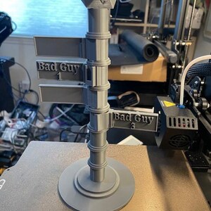 May include: A 3D printed gray signpost with a 20-sided die on top. The signpost has three signs that say "Bad Guy 1", "Bad Guy 2", and "Bad Guy 3".
