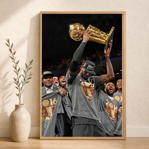 May include: A framed vertical poster featuring a photograph of basketball players celebrating a championship victory. The central figure holds a large gold trophy overhead. Players wear grey t-shirts with 2016 championship graphics and black caps.