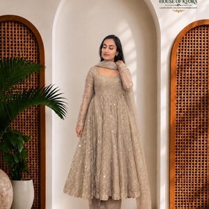 May include: A beige long-sleeved Anarkali suit featuring intricate silver embroidery and sequins throughout the bodice and flared skirt. The ensemble includes matching wide-leg trousers and a sheer dupatta, with the House of Kyora logo visible.
