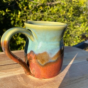 May include: A ceramic mug featuring a smooth, rounded handle and a reactive glaze finish. The upper portion of the mug is a soft sage green, transitioning into a rich, earthy brown and deep orange base with a glossy, multi-toned appearance.