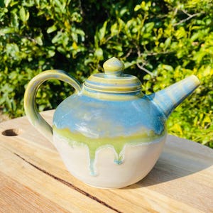 May include: A ceramic teapot featuring a rounded body with a glossy glaze that transitions from a light cream base to a vibrant blue and lime green drip pattern. It includes a matching lid with a small knob, a curved handle, and a tapered spout.