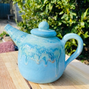 May include: A ceramic teapot with a rounded body, a curved handle, and a spout. The teapot features a light blue glaze with a darker blue drip pattern cascading from the top rim. A matching circular lid with a small knob sits on the top opening.