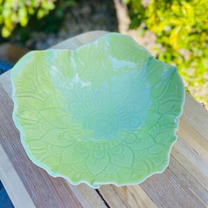 May include: A light green ceramic bowl with a glossy finish and a scalloped, wavy rim. The interior surface features a detailed, embossed mandala-style floral pattern radiating from the centre, creating a textured look across the bowl.