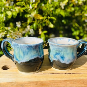 May include: Two ceramic mugs featuring a rounded shape and a handle on the side. The exterior glaze transitions from a deep black base to a vibrant blue and turquoise drip pattern near the rim. The interior of each mug is finished in a smooth white glaze.