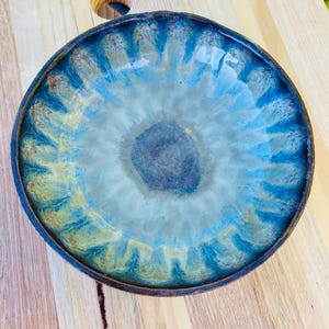 May include: A round ceramic bowl featuring a reactive glaze with a dark blue centre that transitions into lighter shades of turquoise and pale yellow towards the scalloped rim. The bowl has a smooth, glossy finish and a dark, earthy brown edge.