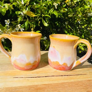 Peut inclure: Deux tasses en céramique présentant une silhouette lisse et arrondie avec une anse sur le côté. L'émail passe d'un jaune chaud au bord à une crème douce au centre, avec un motif de gouttes orange vif et blanc près de la base.