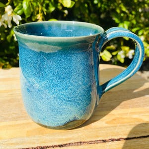 May include: A ceramic mug featuring a speckled blue and teal glaze with a smooth, rounded body and a curved handle. The surface displays a gradient of deep ocean blue at the base transitioning to a lighter turquoise hue near the rim.