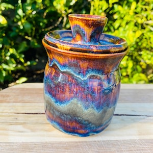 May include: A ceramic butter crock featuring a glossy glaze with swirling bands of deep blue, vibrant purple, and warm amber tones. The container has a rounded body and a matching lid with a central knob handle, showcasing a smooth finish.
