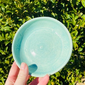 May include: A round, light turquoise ceramic dish featuring a subtle, embossed floral pattern across its interior surface. The bowl has a smooth, glossy glaze finish and a slightly raised rim, showcasing a consistent colour throughout the piece.