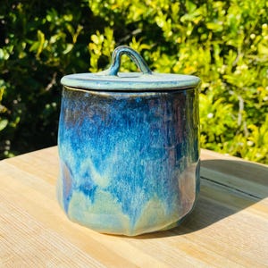 May include: A ceramic jar with a lid featuring a glossy glaze in shades of deep blue, turquoise, and hints of light green. The cylindrical container has a smooth, rounded body and a small, arched handle centred on top of the fitted lid.