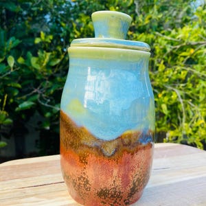 May include: A cylindrical ceramic jar with a matching lid, featuring a smooth glaze that transitions from a light green top to a sky blue middle, and a textured, mottled base in shades of deep orange, rust, and dark brown with a glossy finish.