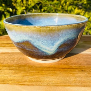 May include: A ceramic bowl featuring a smooth, rounded shape with a reactive glaze finish. The exterior and interior display a blend of deep brown, earthy tones at the base, transitioning into vibrant shades of blue and light turquoise near the rim.