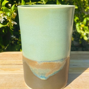 May include: A cylindrical ceramic tumbler featuring a smooth, matte light green glaze on the upper portion and a dark brown glaze on the lower section. The two colours meet in an irregular, organic drip pattern with subtle blue-toned accents.