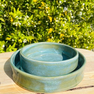 May include: Two round ceramic bowls with a glossy, mottled teal and sage green glaze. The smaller bowl is nested inside the larger, wider bowl. Both pieces feature smooth, curved walls and a consistent, reflective finish across their surfaces.