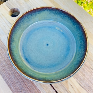 May include: A round ceramic bowl featuring a smooth, glazed finish with a gradient of colours transitioning from a light blue centre to a deep teal and dark brown rim. The bowl has a simple, circular shape with a slightly raised edge and a glossy surface.