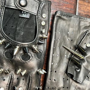 May include: A pair of black fingerless leather gloves featuring silver and black conical metal spikes, circular studs, and clear rhinestones. The gloves have perforated detailing and a snap button closure at the wrist with a size M label visible.