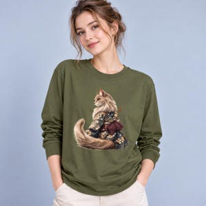 Puede incluir: Una camiseta de manga larga color verde oliva con un estampado gráfico de un gato esponjoso que lleva un kimono oscuro con motivos y un fajín obi granate. El gato se muestra desde atrás, luciendo su larga cola peluda y el diseño floral.