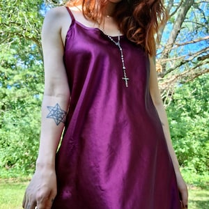 May include: A knee-length slip dress in a deep, lustrous purple satin fabric. The garment features thin spaghetti straps and a simple, fluid silhouette that drapes loosely over the body, showcasing a smooth, reflective surface texture.
