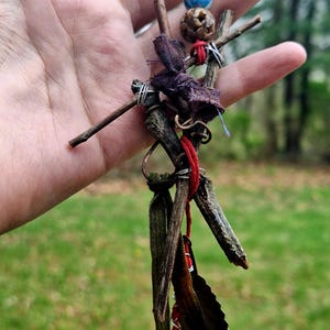 May include: A handmade charm featuring twigs, fabric, and beads. The charm has a red string, a gold-colored medallion, and dark green leaves. The beads are blue, brown, and iridescent. The charm is held in a hand against a blurred green background.