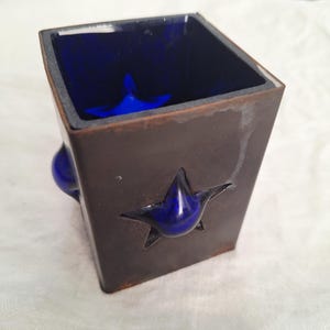 May include: A square, brown ceramic container with a dark blue interior. A blue star shape is cut out of the front, with a blue star shape inside. A blue, rounded shape is on the side. The container is likely for decorative purposes.