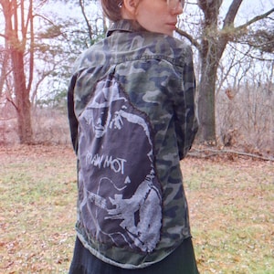 May include: Camouflage-patterned button-up shirt with a large, gray graphic on the back. The graphic appears to be a stylized image with the text "RAW MOT". The shirt is worn over a dark skirt.
