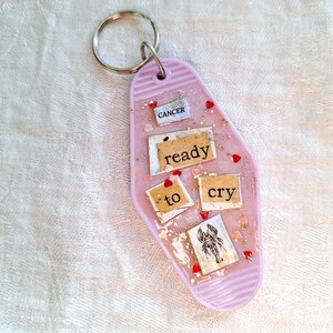 May include: Pink keychain with a light pink resin charm shaped like a motel key. The charm has glitter, gold flakes, red hearts, and paper scraps with the words "ready to cry" and the zodiac sign "Cancer".