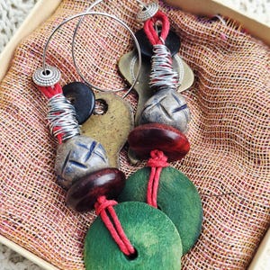 May include: Handmade earrings with silver hoops and a variety of beads. The earrings feature red cord, black and brown wooden beads, and green circular beads. The earrings are displayed in a box with a patterned fabric lining.
