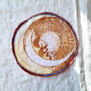 May include: A round, white fabric patch with a brown crescent moon design. The patch is surrounded by a red, blue, and yellow stitched border.