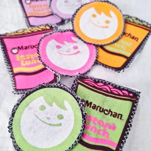 May include: A collection of colorful, handmade fabric patches featuring the text "Maruchan Instant Lunch" and a smiling cartoon character. The patches are in shades of pink, orange, and green, with black stitched edges.