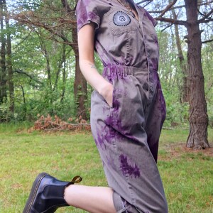 May include: A gray jumpsuit with purple tie-dye accents. The short-sleeved jumpsuit features a button-up front, an elastic waist, and a patch on the chest. The person is wearing black ankle boots with yellow accents.