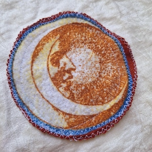 May include: A round, white fabric patch with a brown crescent moon design. The patch is surrounded by a red and blue stitched border.