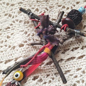 May include: A handcrafted charm featuring twigs, fabric scraps, and beads. The charm includes a yellow eye-shaped bead, a tassel, and a mix of natural and colorful elements. The design incorporates red, purple, and yellow accents, with silver wire detailing.