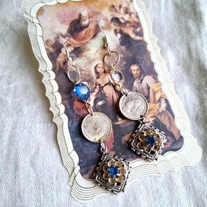 May include: A pair of silver earrings with blue and gold accents. Each earring features a small blue gemstone, a coin, and a square charm with a blue gemstone in the center.