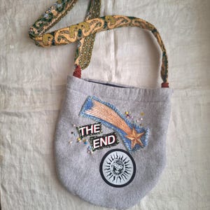 May include: Gray shoulder bag with a patterned strap and the words "THE END" on the front. The bag features a shooting star and sun graphic, with decorative beads. The strap is a mix of green, yellow, and brown patterns.