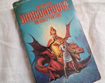 May include: A vintage paperback book titled "Dragonrouge: An Adult Fantasy" by Lin Carter. The cover features a red dragon, a woman riding the dragon, and a warrior. The background is blue.
